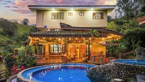 Una casa con piscina, un espacio exterior cubierto con mesas y sillas, y un jardín exuberante.
