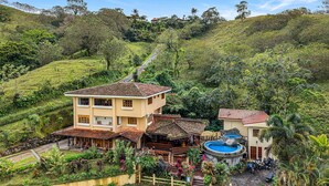 Un edificio de dos pisos con techo de tejas, rodeado de exuberante vegetación y una pequeña piscina.