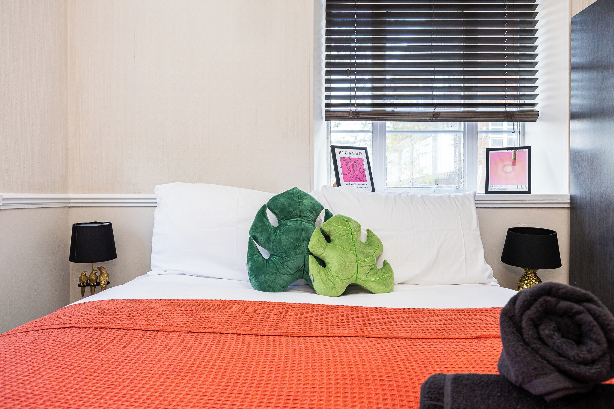 A hotel room with a bed, two pillows, a window with blinds, a lamp, and a framed picture on the wall.