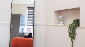 Una habitación con un espejo que refleja una ventana, una pared blanca, una estantería con una escultura de busto y una planta colgante.