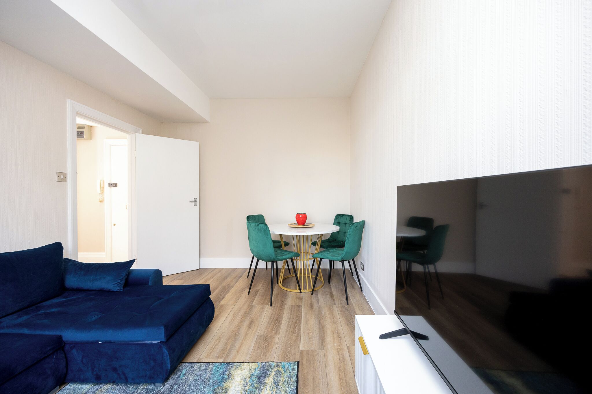 A compact living space with a blue sofa, a small round dining table with green chairs, and a flat-screen TV mounted on the wall.