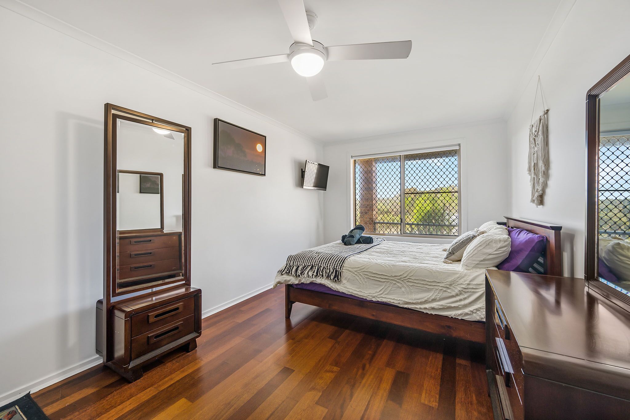 A bedroom with a wooden bed, a dresser, a ceiling fan, a TV, and a window with a view of greenery.
