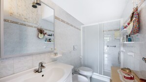 Baño con lavabo, inodoro y ducha. Hay repisas con artículos de aseo y un espejo.