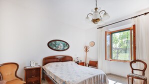 Un dormitorio con una cama, una silla de madera, una mesita de noche, un aparador, un cuadro en la pared y una ventana con vistas a la vegetación.