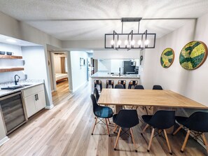 Une cuisine moderne dotée d’un îlot central, un coin repas avec une table et des chaises en bois, et un couloir menant à une chambre.