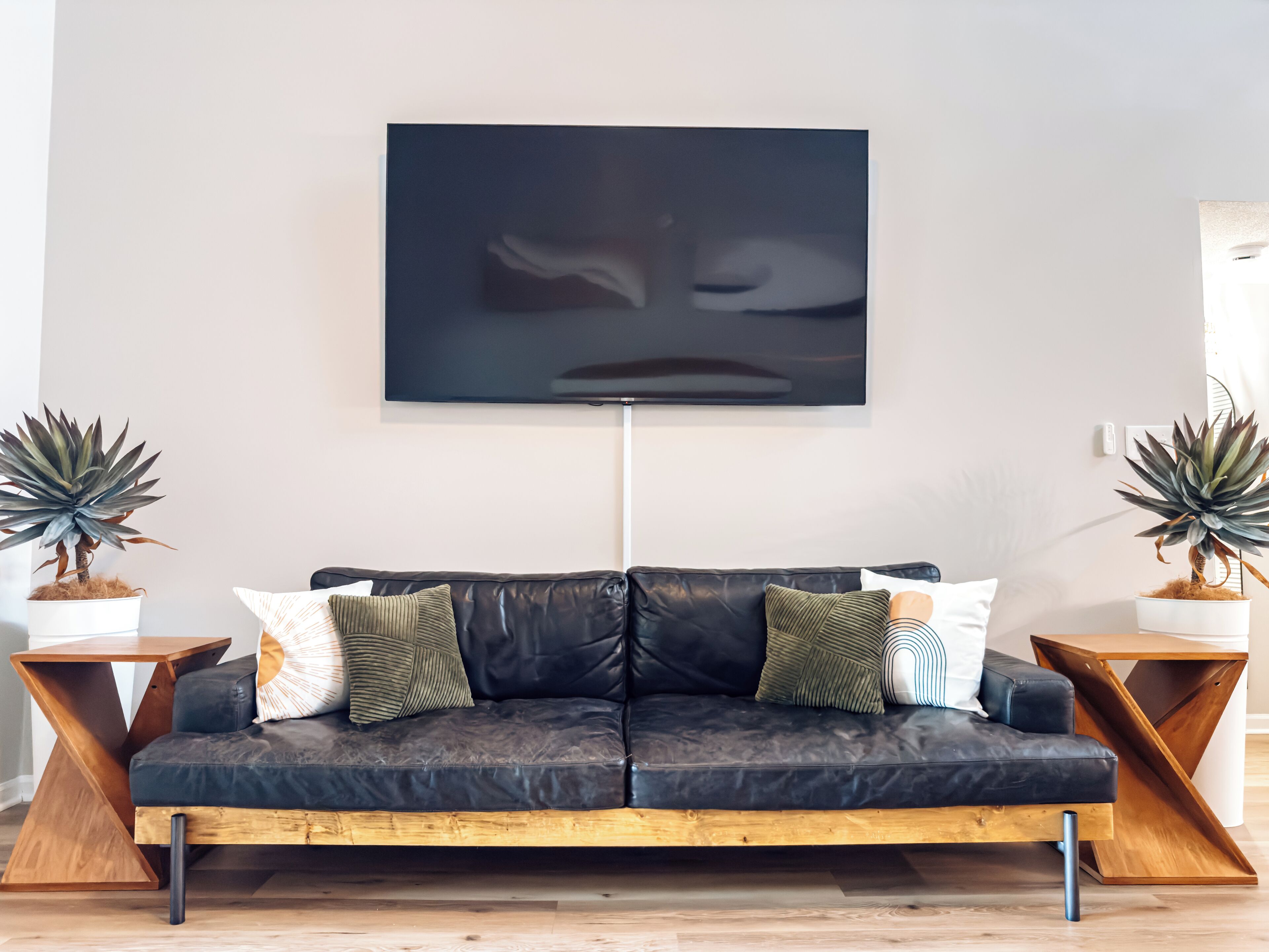 A modern living room with a black leather sofa, two wooden side tables, and two potted plants.