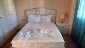 Ein ordentlich bezogenes Bett mit blumigen Kissen und Handtüchern, ein Nachttisch mit Lampe und ein Kleiderschrank mit Schiebetüren.