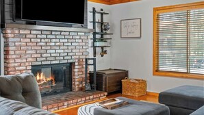 Ein Wohnzimmer mit Kamin aus Backstein, einem darüber montierten Fernseher und einem Fenster mit Jalousien.