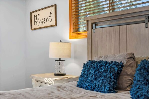 A bedroom with a bed, a nightstand, a lamp, a wall sign, and a window with blinds.