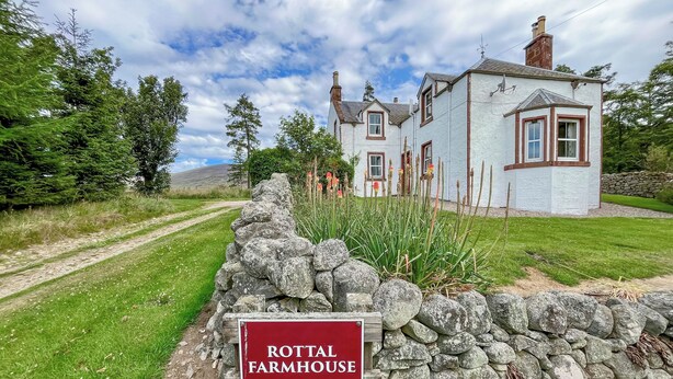 A stone wall with a sign reading "ROTTAL FARMHOUSE" in front of a white house with brown trim and multiple windows.