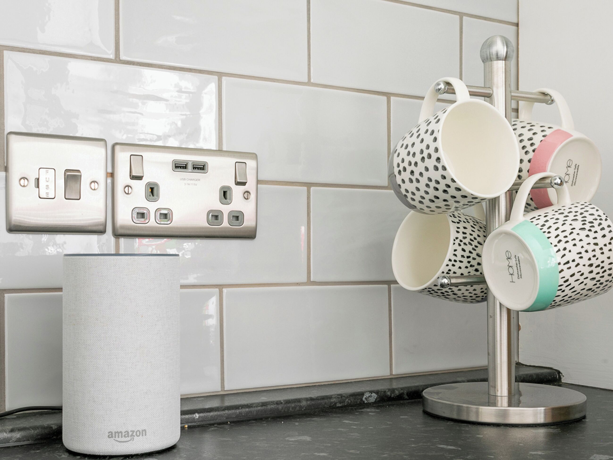 A kitchen with a white tiled backsplash, a wall-mounted USB charging station, and a mug holder with variously patterned mugs.