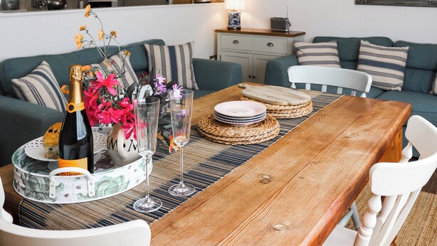 Un coin repas avec une table en bois, des chaises blanches et un plateau de service contenant des assiettes et une bouteille de champagne.