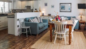 Une cuisine moderne avec des meubles blancs, un coin repas doté d’une table et de chaises en bois, et un salon avec un canapé bleu et des coussins à rayures.