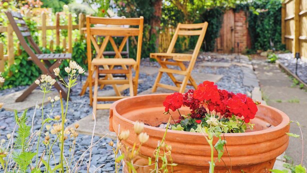A garden with a terracotta pot of red flowers, wooden chairs, and a wooden fence. - 2 bedroom accommodation in Cheltenham (Cheltenham)