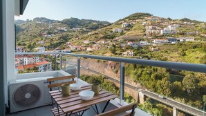 Un balcon équipé d’une table, de chaises et d’un ventilateur. Le balcon offre une vue sur un paysage vallonné parsemé de bâtiments.
