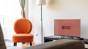 Une chambre d’hôtel moderne avec un seul fauteuil orange, un téléviseur à écran plat affichant le message « PRÊT POUR L’ARRIVÉE DU CLIENT » et une lampe de sol.