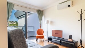 Un salon moderne avec un canapé gris, un fauteuil orange, un téléviseur à écran plat et un balcon offrant une vue sur une colline.