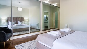Una habitación de hotel moderna con una cama grande, una zona de estar separada, un vestidor y un baño visible a través de una puerta de cristal.