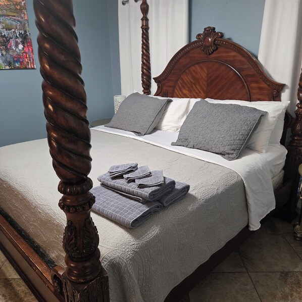 A four-poster bed with a white bedspread and grey pillows, a folded grey towel, and a decorative headboard.