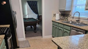 A kitchen with green cabinets, a granite countertop, a dishwasher, and a pool table in the background. - GameDay! University of South Alabama Poolside Getaway🇺🇲Veteran Own ⚓️ (Mobile)