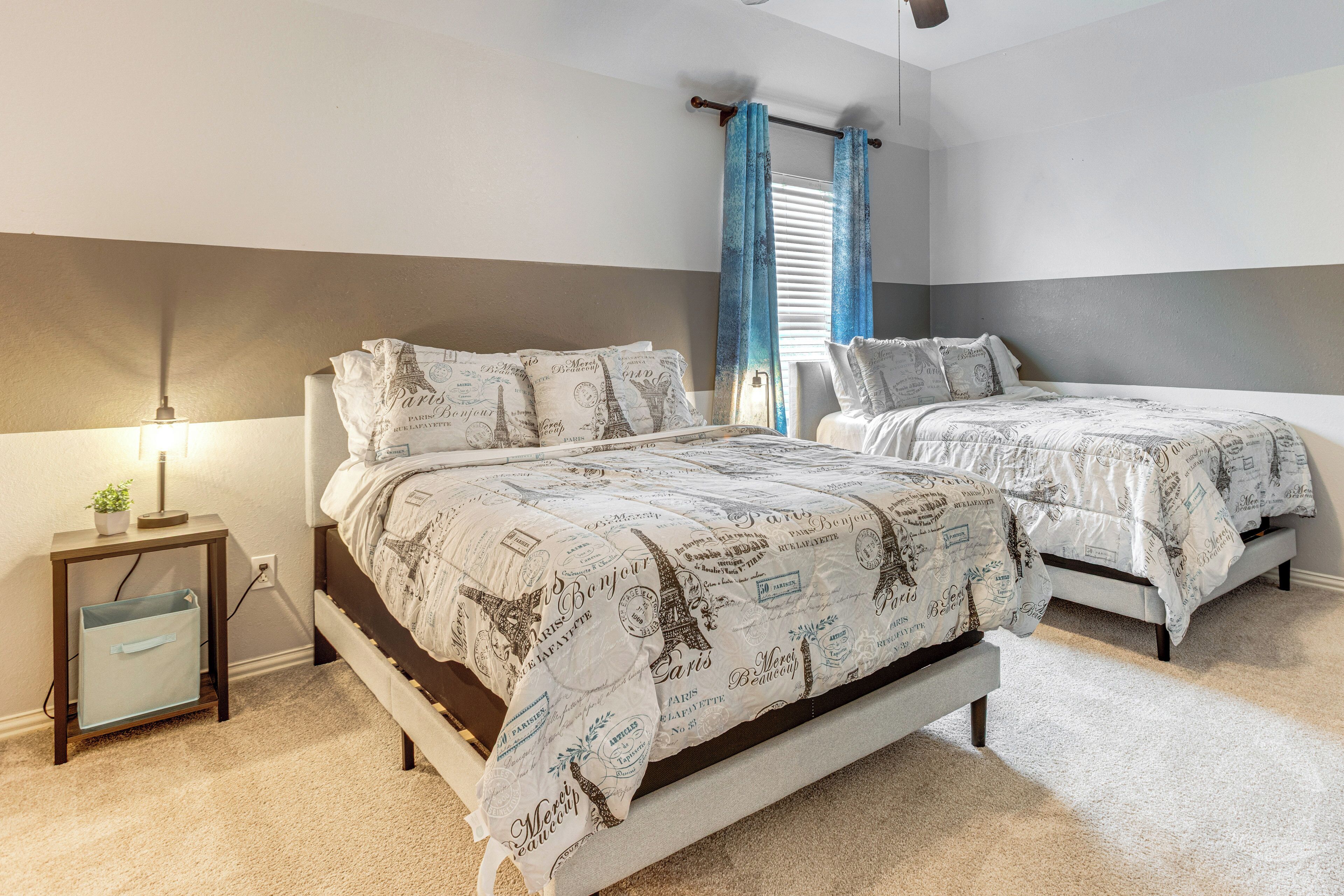 Quarto com duas camas, mesa de cabeceira, luminária e janela com cortinas.