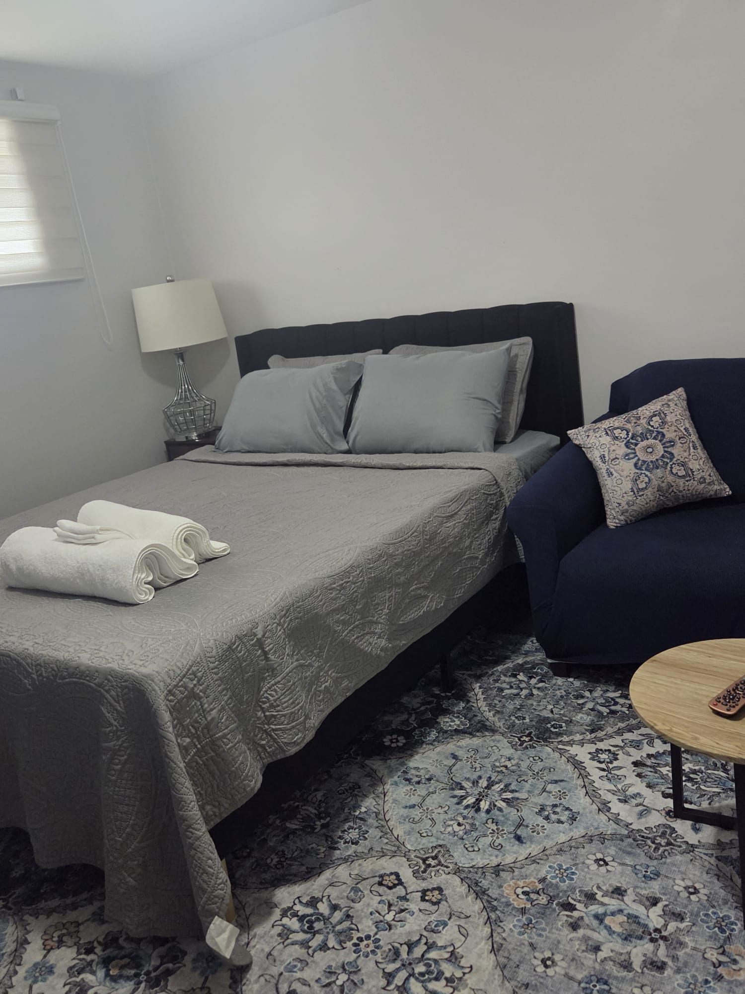 A single bed with a grey bedspread, a bedside table with a lamp, a blue armchair, and a small round table with a remote control.