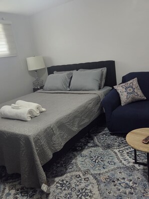 Ein Einzelbett mit grauer Bettdecke, ein Nachttisch mit Lampe, ein blauer Sessel und ein kleiner runder Tisch mit Fernbedienung.