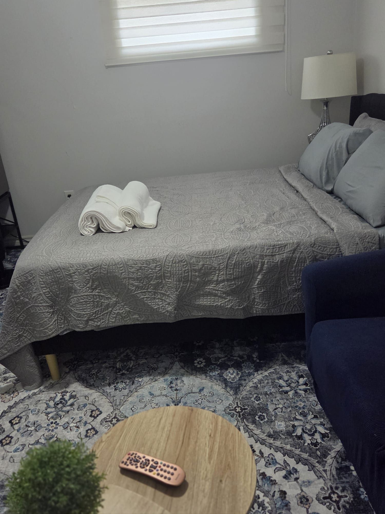 A neatly made bed with a folded towel, a remote control on a round wooden table, and a blue sofa in a room with patterned carpet.