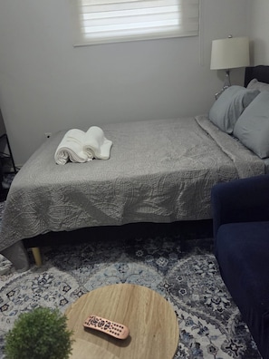 Un lit bien fait, avec une serviette pliée, une télécommande posée sur une petite table ronde en bois, et un canapé bleu dans une pièce dont le sol est recouvert d’une moquette à motifs.