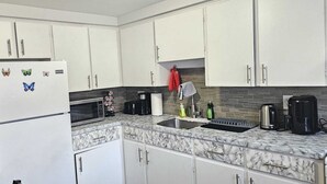 Cocina compacta con armarios blancos, encimera de mármol, refrigerador con imanes, cafetera y hervidor.