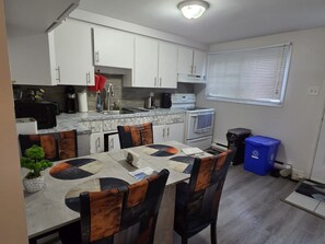 Una cocina compacta con armarios blancos, un microondas, una estufa y un lavamanos. Hay una mesa de comedor con sillas y un cubo de basura azul.