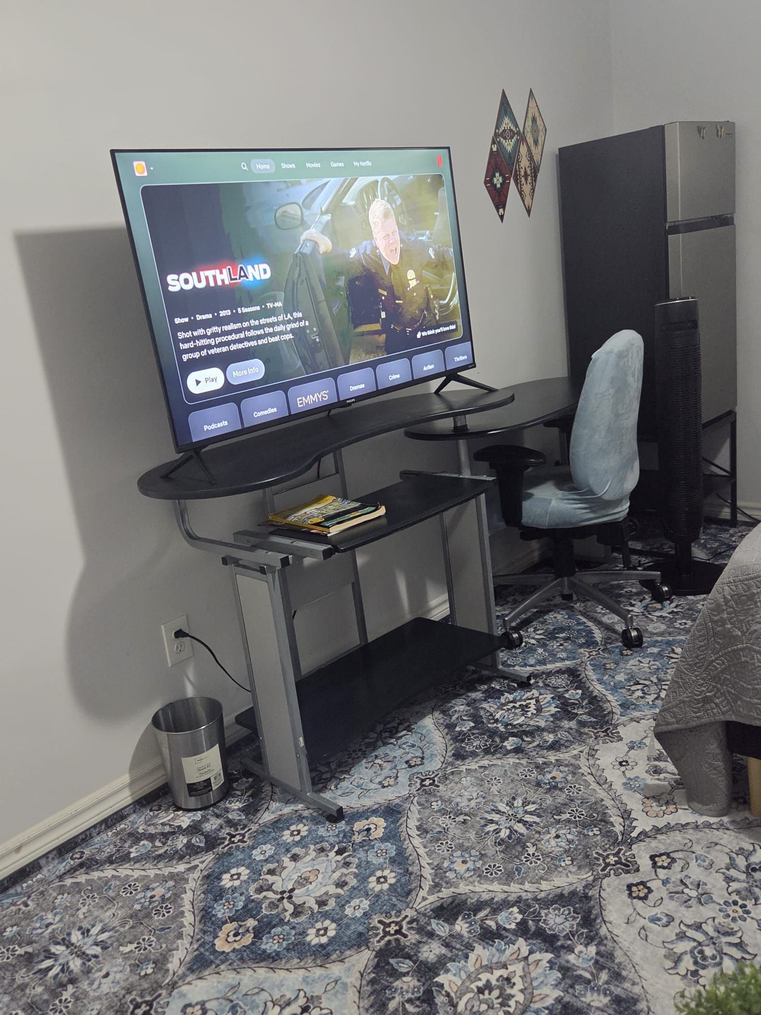 A television mounted on a wall with a remote control, a desk with a chair, a rug, and a small trash can.