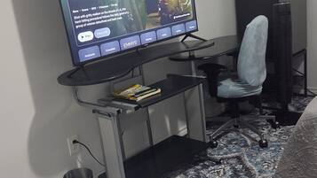 A television mounted on a wall with a remote control, a desk with a chair, a rug, and a small trash can.