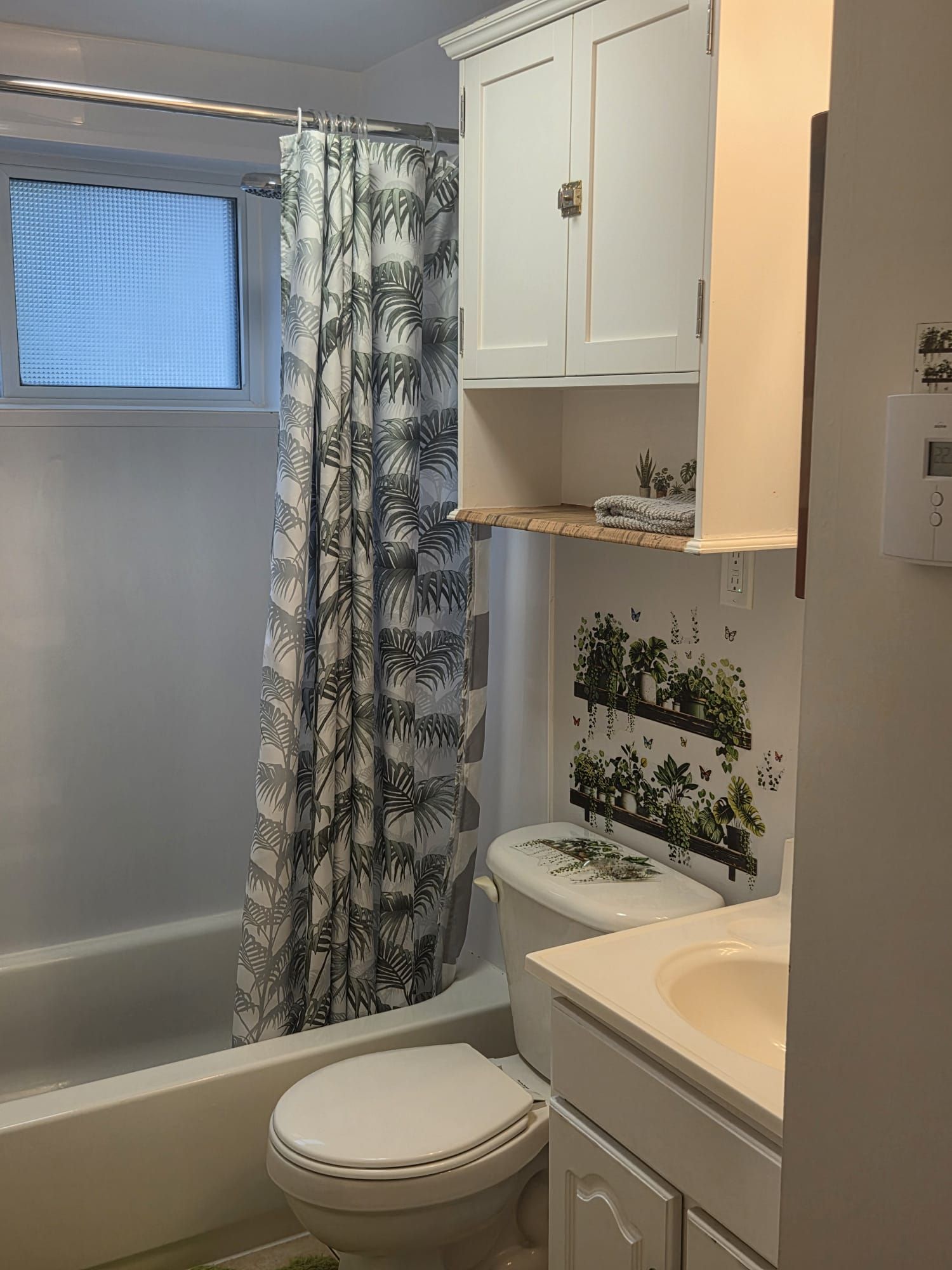 Une salle de bain avec des toilettes, un rideau de douche orné d’un motif de feuilles, une fenêtre et une étagère murale avec des plantes.