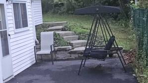 Un espace patio avec une chaise suspendue et une petite table, entouré d’un mur de pierre et d’une clôture verte.