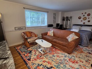 Una sala de estar amoblada con un sofá marrón, un sillón estampado, una mesita blanca pequeña y una alfombra colorida.