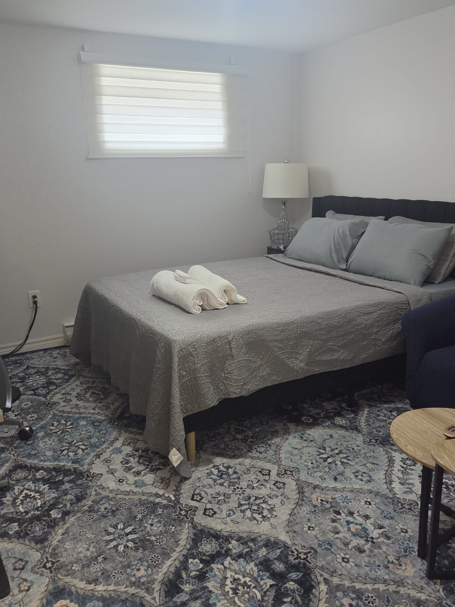 A hotel room with a bed, a bedside lamp, a window with blinds, a patterned carpet, and a small table.