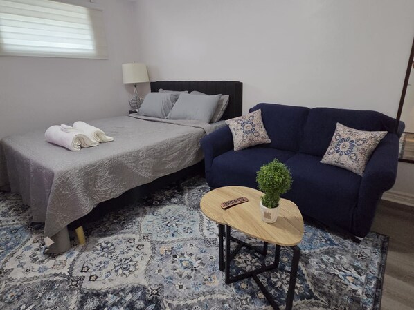 Un dormitorio con una cama, un sofá azul, una mesita con una planta y una alfombra estampada.
