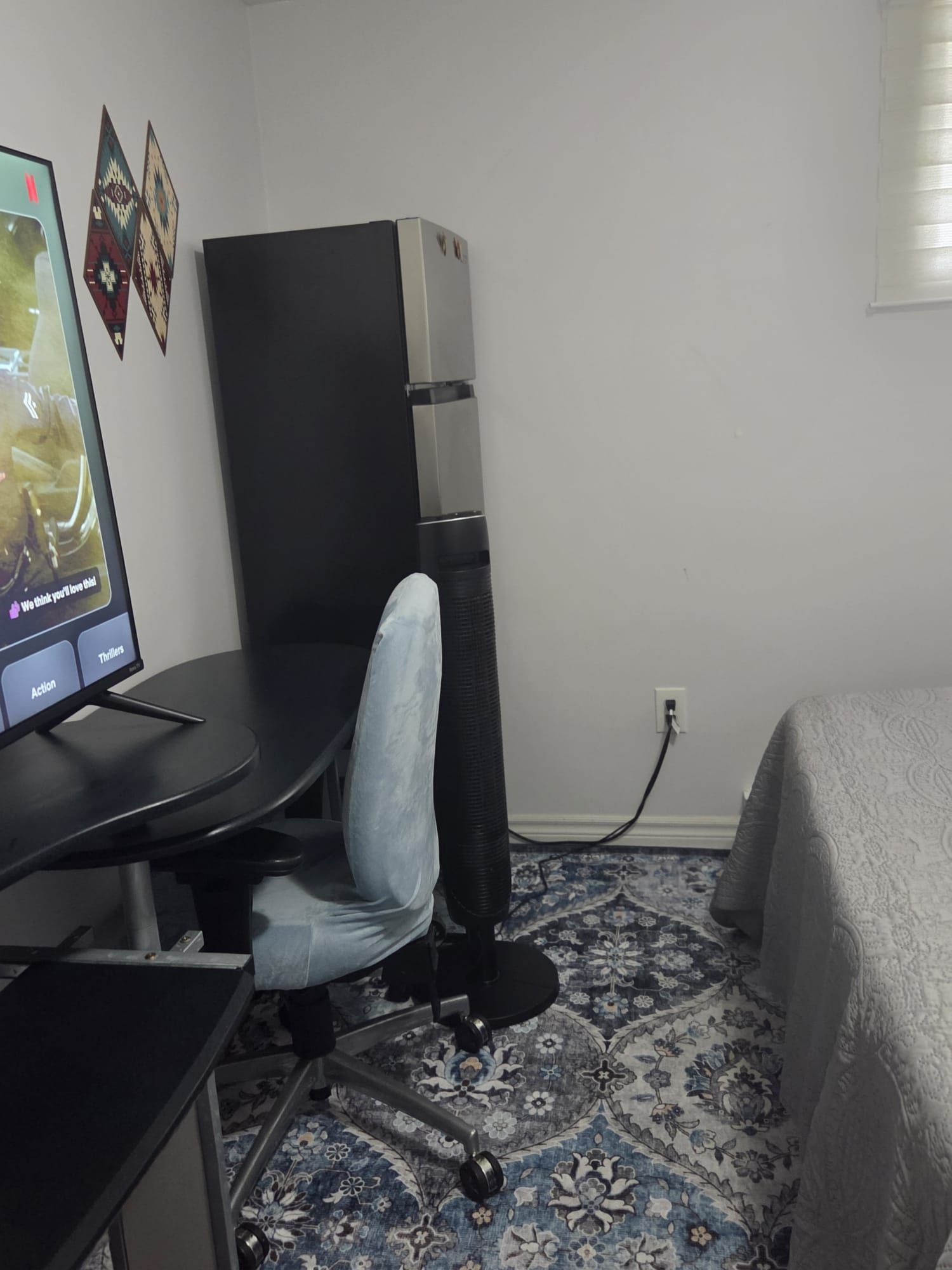 A room with a desk, a computer, a chair, a standing fan, and a bed with a patterned bedspread.