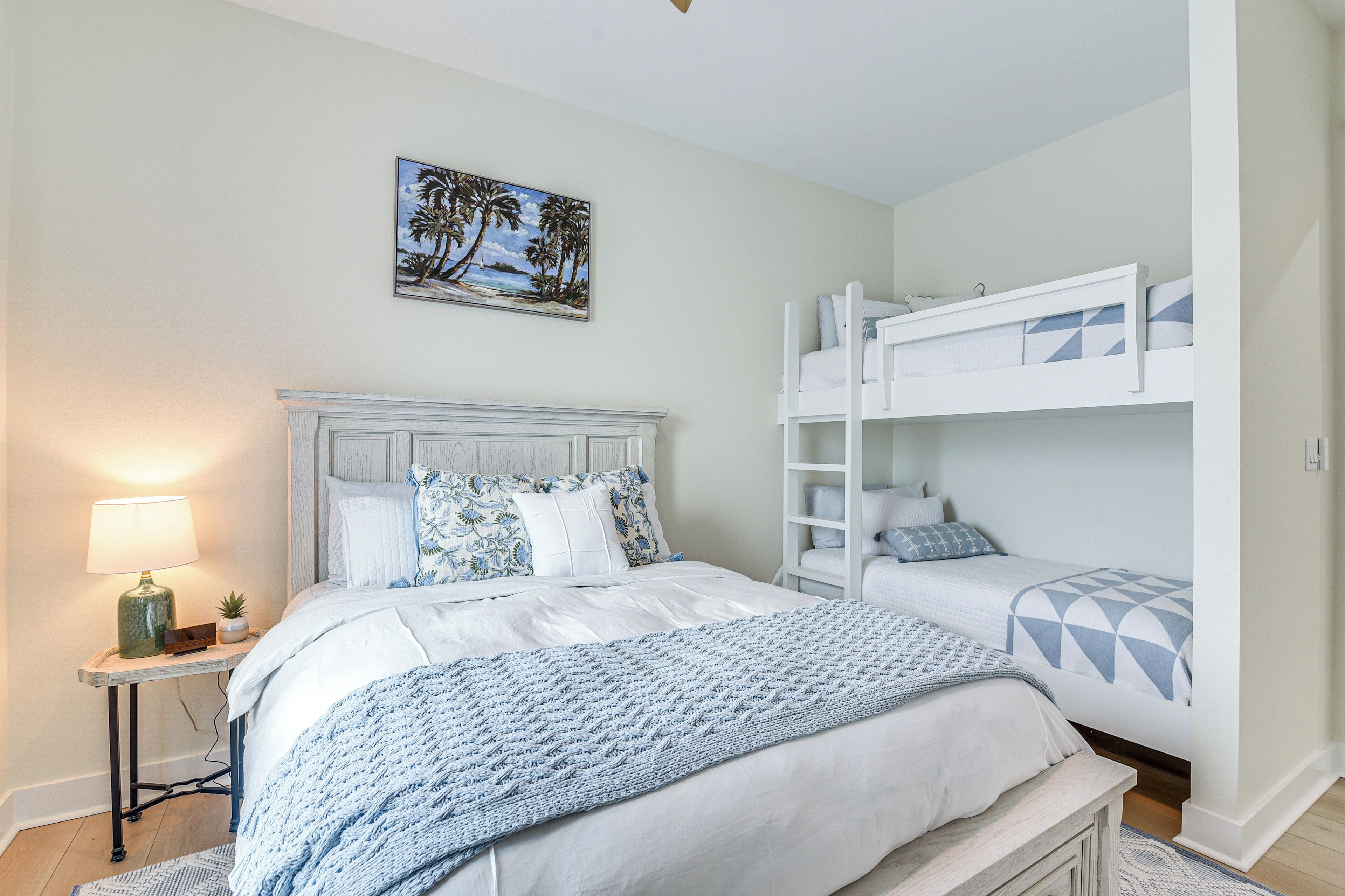 Une chambre avec un grand lit, une table de chevet, une lampe, un tableau représentant des palmiers et un lit superposé.