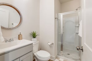 Un baño con un lavamanos blanco, un espejo redondo, un inodoro y una cabina de ducha con mampara de vidrio.