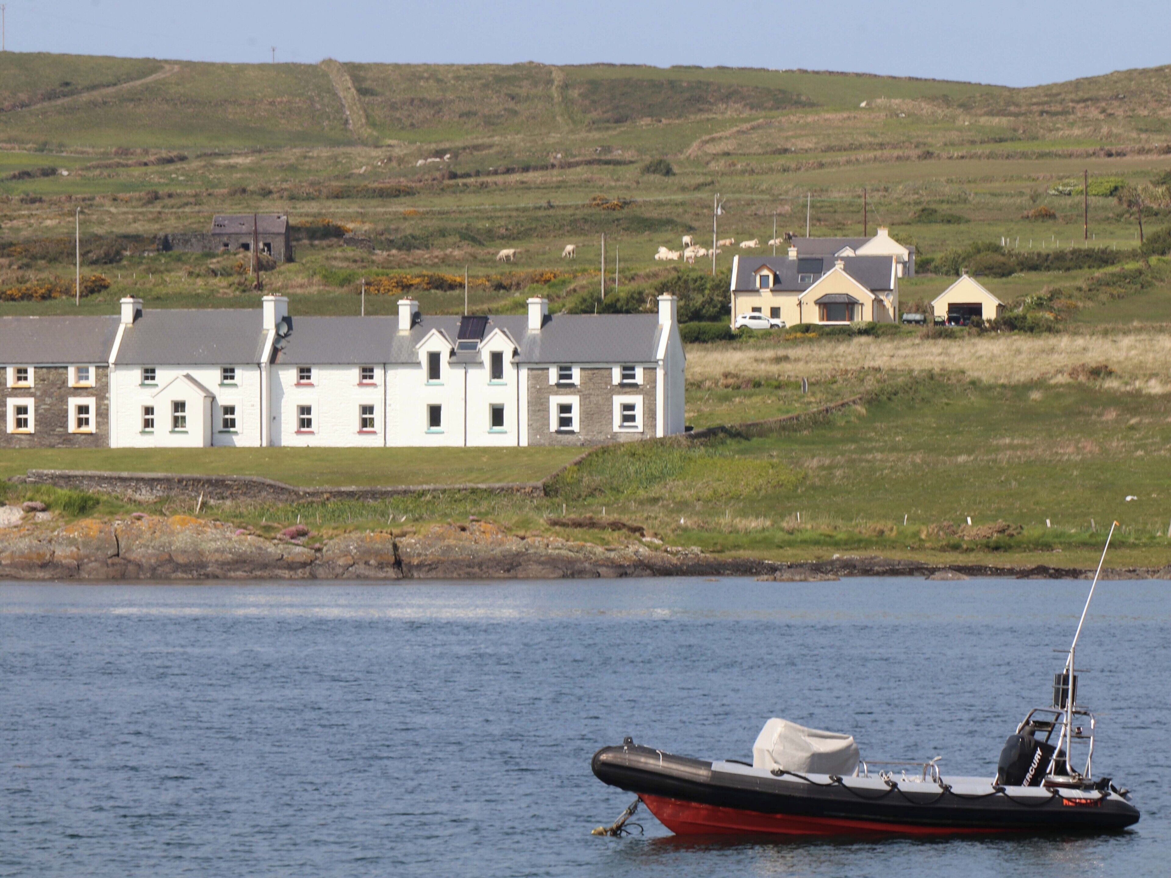 A coastal scene with a row of white houses with grey roofs, a small boat anchored near the shore, and a grassy hill in the background.