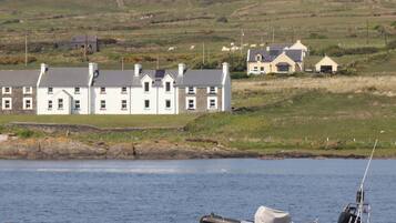 A coastal scene with a row of white houses with grey roofs, a small boat anchored near the shore, and a grassy hill in the background.