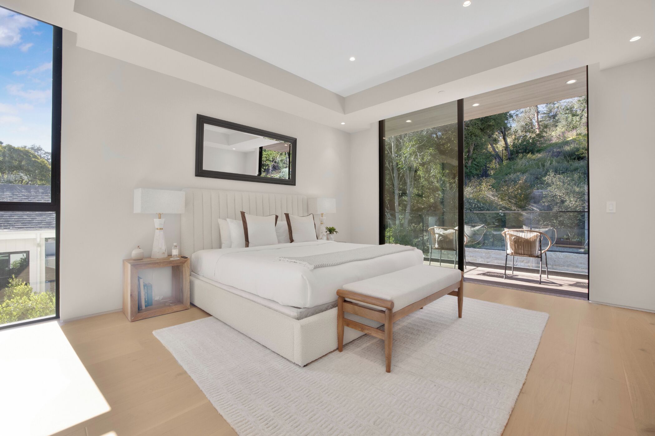 A modern bedroom with a large bed, a bedside table, a mirror, and a view of a balcony.