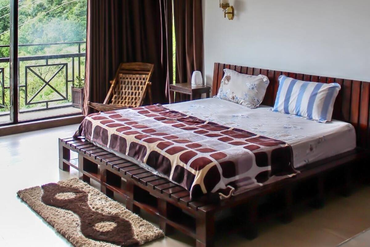 Ein Schlafzimmer mit einem großen Bett, einer hölzernen Bank, einem Nachttisch und Blick auf Grün hinter dem Fenster.