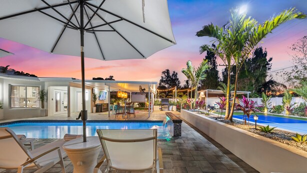 A poolside area with lounge chairs and an umbrella, surrounded by a landscaped garden and a pool. - Wellness: Pool-Sauna-HotTub-ColdPlunge-Pickleball (Bradenton)