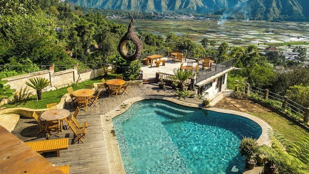 A pool area with wooden furniture, a hanging chair, and a view of mountains. - HK Villa's (Sembalunbumbung)