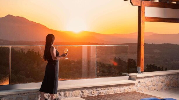 Een vrouw staat op een balkon, genietend van een glas wijn met uitzicht op een zonsondergang en bergen.