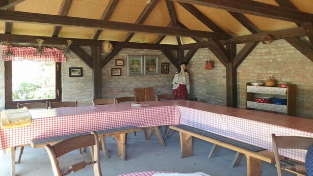 A rustic dining area with a long table covered in a checkered tablecloth, wooden benches, and a brick wall with framed pictures.