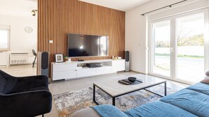 A modern living room with a large flat-screen TV, wooden panel wall, white entertainment unit, and a large sliding glass door to the outdoors. - Holiday Home Iva - Three Bedroom Holiday Home (Šibenik)